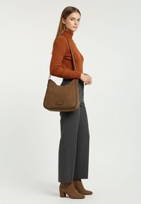 Braune Umhängetasche aus Wildleder mit geflochtenem Detail entlang des Randes, mit flachem Boden. Das Model trägt einen rostfarbenen Rollkragenpullover und graue Hosen.