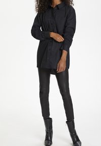 Sort skjorte med lange ærmer, der har en brystlomme og buet kant, parret med sorte tætsiddende leggings og ankelstøvler.