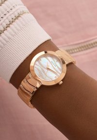 Reloj de oro rosa con una esfera de nácar. Presenta un diseño ondulado, acentos de cristal y una pulsera elegante, llevado en una muñeca.