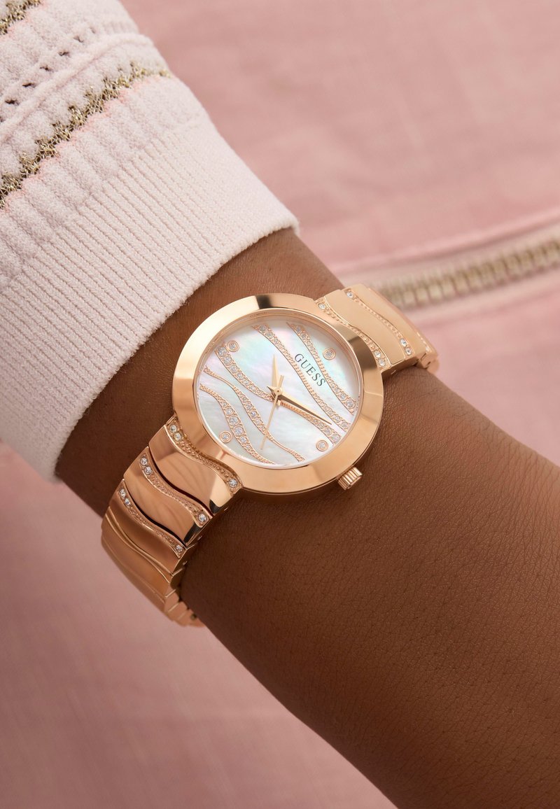 Reloj de oro rosa con una esfera de nácar. Presenta un diseño ondulado, acentos de cristal y una pulsera elegante, llevado en una muñeca.