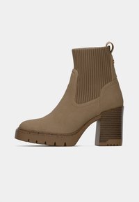 Botine maro din suede, cu turul din material ribetat, toc gros și talpă zimțată. Include o bucată de prindere pentru ușurința la îmbrăcare și accente texturate.