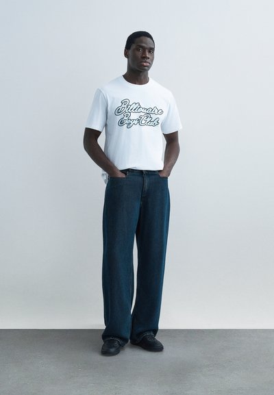 Άνδρας που στέκεται με τα χέρια στις τσέπες, φοράει λευκό μπλουζάκι με τύπωμα, σκούρα τζιν φαρδιάς γραμμής και μαύρα παπούτσια, μπροστά από ένα απλό φωτεινό φόντο.