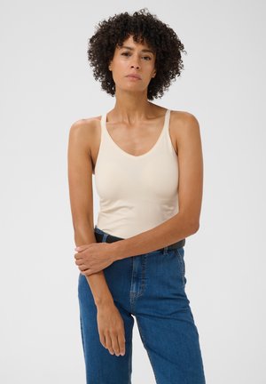 Femme aux cheveux bouclés portant un débardeur beige et un jean bleu, debout les bras croisés devant un fond clair uni.