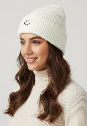 White ribbed knit beanie featuring a circular black smiley face patch. Soft texture, fitted design, and slight slouch for casual wear.