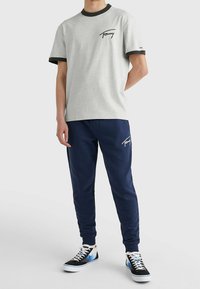 Grå t-shirt med kort ärm och svarta detaljer, med en logo på bröstet, ihop med marinblå träningsbyxor och svarta sneakers med blå detaljer.