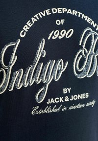 Tissu bleu marine avec texte blanc indiquant "Département Créatif de 1990, Indigo par Jack & Jones, Fondé en mille neuf cent quatre-vingt-dix."
