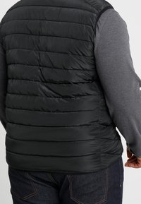 Czarna pikowana kamizelka z poziomym wzorem, lekki materiał i gładka tekstura; noszona na szarej koszuli z długim rękawem.