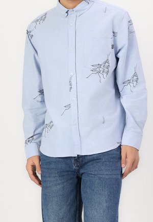Light blue button-up shirt with black praying mantis insect prints worn by a person with blue jeans, standing against a plain background.
