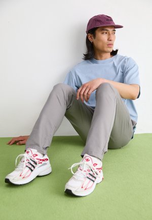 Jeune homme portant une casquette violette, un t-shirt bleu clair, un pantalon gris et des baskets blanches, assis sur un sol vert contre un mur blanc, regardant vers la droite.