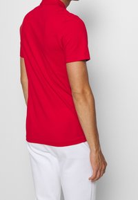 Red short-sleeve polo shirt made of smooth fabric, featuring a classic collar and a fitted design, worn with white pants.