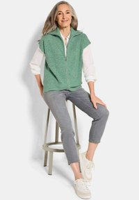 Gilet en tricot vert avec fermeture éclair, porté sur une chemise blanche. Pantalons cropped gris et baskets claires. Modèle assis sur un tabouret de couleur claire.