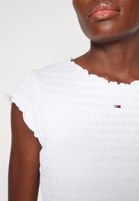 Personne à la peau foncée portant un haut blanc texturé à manches courtes avec un petit logo Tommy Hilfiger sur la poitrine.