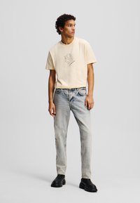 T-shirt à manches courtes de couleur jaune clair avec un motif graphique, associé à un jean bleu délavé et des bottes noires à la cheville. Texture du tissu lisse.