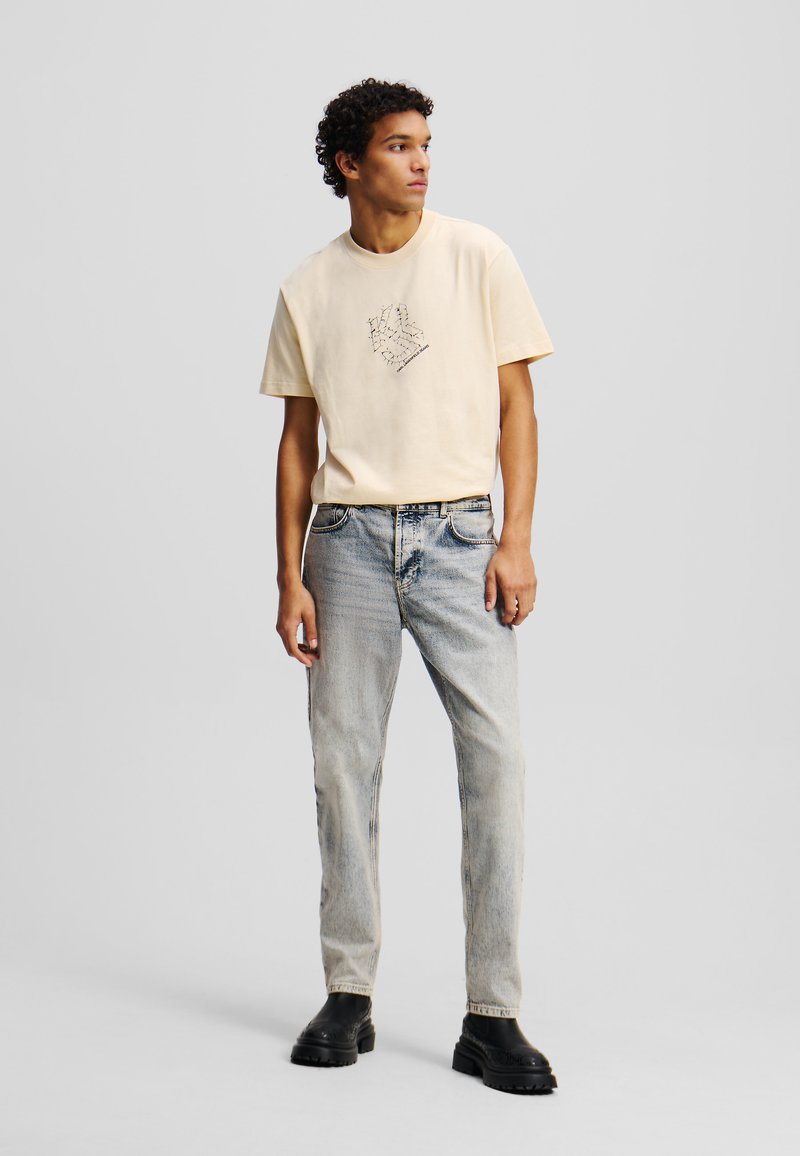 T-shirt à manches courtes de couleur jaune clair avec un motif graphique, associé à un jean bleu délavé et des bottes noires à la cheville. Texture du tissu lisse.