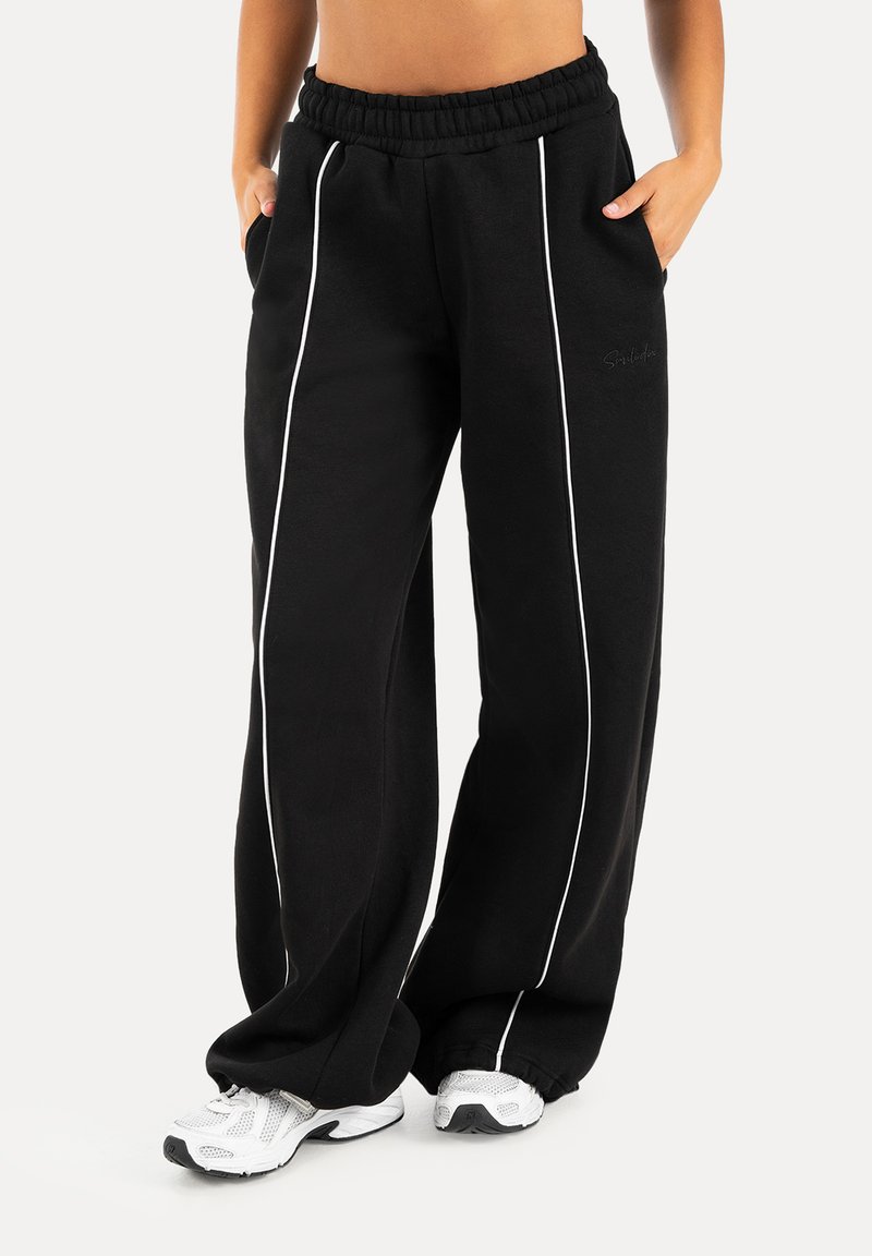 Pantaloni della tuta neri a gamba larga con righe verticali bianche indossati con sneakers bianche, mani del modello nelle tasche e top corto visibile.