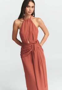Ruda spalva halter stiliaus suknelė su surinkta apykakle, rakto skylutės iškirpte ir drapiruotėmis, su juosmeniu surišta, pagaminta iš lengvo audinio.