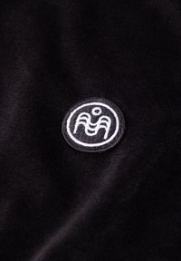 Must full must veluurne tekstuur, millel on paremas alanurgas ringikujuline valge tikandiga logo, mis esitleb stiliseeritud disaini.