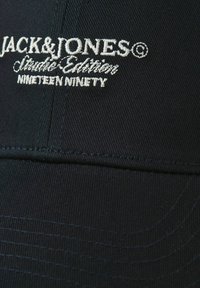 Juodas audinio kepurė su balta siuvinėta užrašu „JACK & JONES Studio Edition Nineteen Ninety“ priekinėje panelėje.