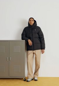Schwarze Pufferjacke mit Kapuze, Reißverschluss und einem Logo. Beigefarbene Hose und graue Turnschuhe. Grauer Spind im Hintergrund.