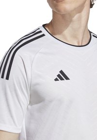 Maglietta sportiva bianca con strisce nere diagonali sulle spalle e un logo Adidas nero. Il tessuto presenta un motivo a chevron testurizzato.