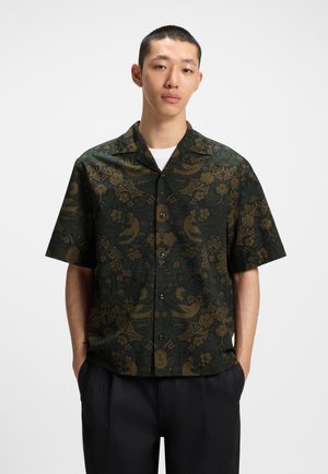 Korte mouwen shirt van donker groene stof met een bloemen- en vogelpatroon in goud. Het heeft een knoopsluiting aan de voorkant en een uitgespreide kraag.