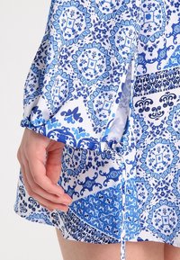 Indumento a fantasia blu e bianco con disegni floreali e geometrici, maniche arricciate e un dettaglio con cordoncino ai polsini.