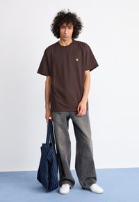 T-shirt oversize marrone con un piccolo logo giallo, abbinato a jeans grigi larghi e a una borsa tote blu navy con strisce bianche. Sneakers bianche.