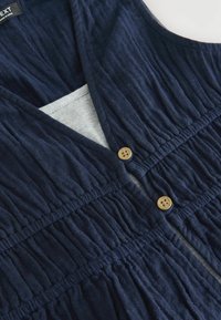 Robe sans manches bleu marine avec un décolleté en V, un texture froncée et deux boutons en bois à l'avant. Doublure grise claire visible à l'intérieur.