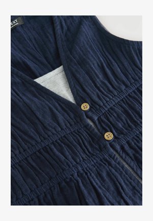 Mornarsko modra brezrokavna obleka z v-izrezom, nabranimi teksturami in dvema lesenima gumboma spredaj. Podloga v svetlo sivi barvi vidna znotraj.