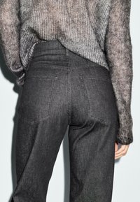 Persona che indossa jeans in denim nero a vita alta e un maglione grigio lavorato a maglia, visto di spalle dalla vita alle cosce su uno sfondo neutro.