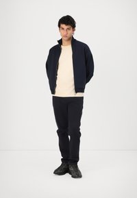 Marine bomberjakke over en beige t-skjorte, kombinert med mørke bukser og svarte joggesko. Stoffet ser glatt ut, med en strukturert design.