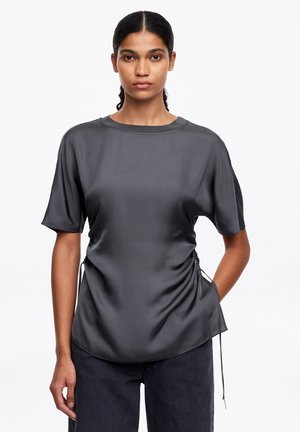 Frau mit dunklem Haar im Pferdeschwanz, die eine kurzärmelige, dunkelgraue Satinbluse mit seitlichen Kordelzügen und schwarze Hosen trägt.