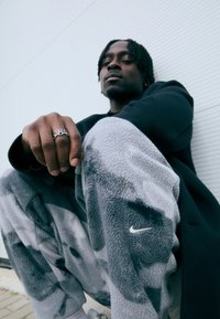 Fekete dzsekit és szürke fleece Nike nadrágot viselő férfi ül egy fehér falnak támaszkodva, ezüstgyűrűs keze a térdén pihen.