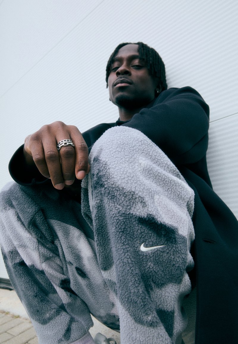 Fekete dzsekit és szürke fleece Nike nadrágot viselő férfi ül egy fehér falnak támaszkodva, ezüstgyűrűs keze a térdén pihen.