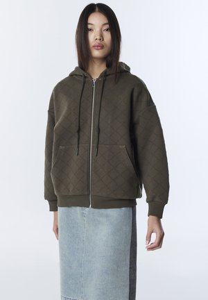 Junge Frau trägt eine gesteppte dunkelgrüne Kapuzenjacke mit Fronttaschen und Reißverschluss, kombiniert mit einem hellen Jeans-Midirock, steht vor weißem Hintergrund.