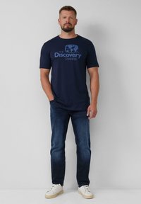 Donkerblauwe T-shirt met korte mouwen met het "Discovery Channel" logo in lichtblauw, gecombineerd met donkere denim jeans en witte sneakers.