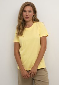 T-shirt en coton jaune avec un col rond et des manches courtes, présenté sur un fond neutre, associé à un pantalon beige.