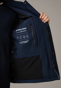 Mano che apre la giacca Strellson Flex Cross blu navy mostrando l'etichetta interna, la tasca con cerniera e le icone informative sul tessuto.