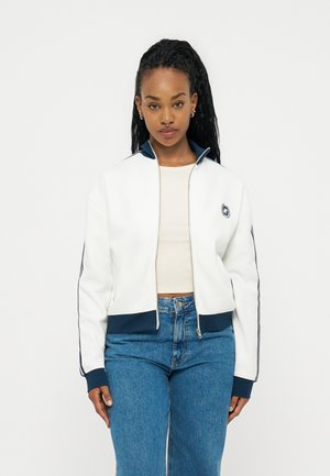 Νεαρή γυναίκα με μακριά πλεξούδα, που φοράει λευκό μπουφάν με φερμουάρ και σκούρη μπλε επένδυση, μπεζ crop top και μπλε τζιν, στέκεται μπροστά σε απλό φόντο.