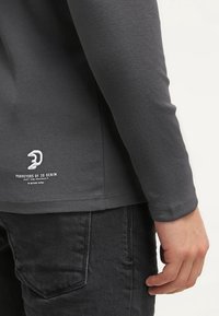 Gray long-sleeve shirt with a fitted design, round hem, and printed logo detail near the bottom. Fabric appears smooth and lightweight.