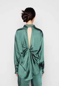 Top de satén verde esmeralda con mangas largas, detalle de volantes en la parte trasera y abertura en forma de llave. La textura suave y la caída fluida realzan el diseño.