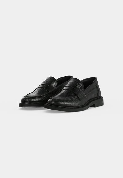 Ζευγάρι μαύρα πλεκτά δερμάτινα loafers με χαμηλά τακούνια, στρογγυλεμένα δάχτυλα και λεπτομέρεια με λουράκι μπροστά σε απλό φωτεινό φόντο.
