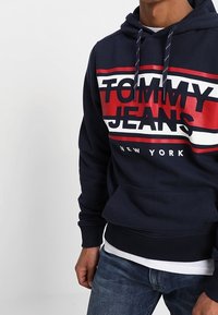 Sudadera de color azul marino con "TOMMY JEANS" y "NEW YORK" impresos en letras rojas y blancas en negrita. Cuenta con cordones y un bolsillo delantero.