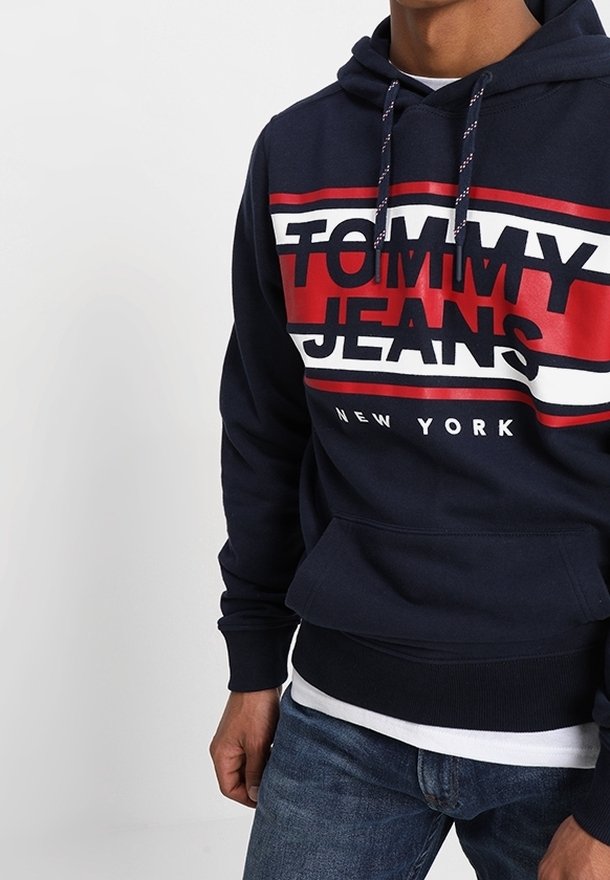 Sudadera de color azul marino con "TOMMY JEANS" y "NEW YORK" impresos en letras rojas y blancas en negrita. Cuenta con cordones y un bolsillo delantero.