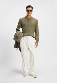 Olivgrüner strukturierter Strickpullover, weiße plissierte Hose, graue Jacke über dem Arm, grau-weiße Sneaker, schwarze Sonnenbrille.