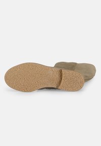 Une seule botte en daim taupe est posée à plat, la semelle en caoutchouc texturée tournée vers le haut, montrant le motif de grip et une légère élévation du talon.