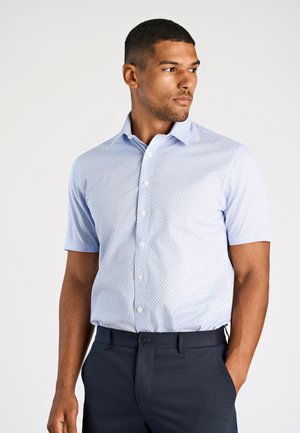 Camicia a maniche corte con bottoni di colore blu chiaro con piccoli pois blu scuro. Presenta un colletto classico e un design aderente. Indossata con pantaloni scuri.