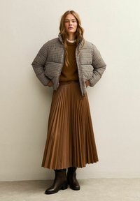 Gekapte houndstooth pufferjack in zwart en bruin, bruine trui, geplisseerde bruine midi-rok en bruine haklaarzen.
