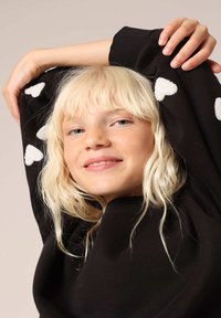 Zwarte trui met witte hartpatronen op de mouwen, ronde halslijn, geribbelde manchetten, gedragen door een kind met lichtblond haar en een ontspannen houding.