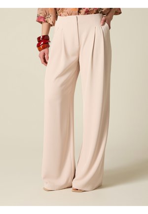 Kvinde iført beige bredbenede bukser, blomsterbluse stoppet ind, stående med den ene hånd i lommen og iført chunky røde armbånd på venstre håndled.